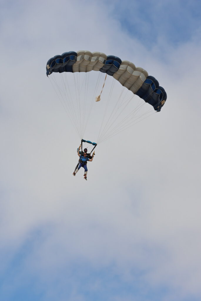 Tandem pair glides high under a blue and white parachute canopy against a cloudy sky